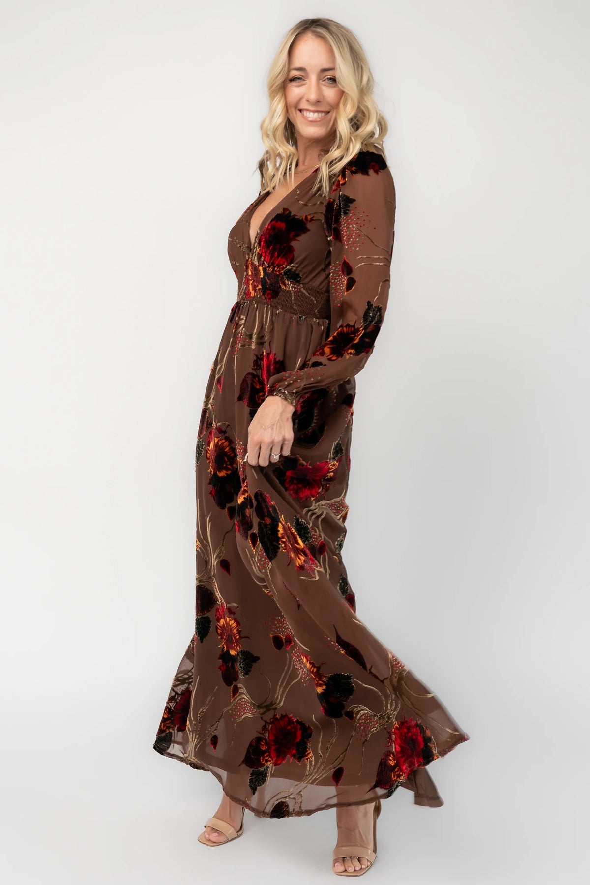 Brown burnout velvet maxi dress with a deep V-neckline, long sleeves, smocked waistband, and flowing asymmetrical hem with gold and burgundy floral accents.