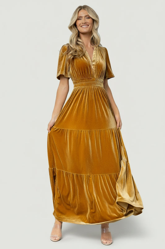 Front view of Artemis Dark Gold velvet maxi dress on model, showing plunging V-neck, short flutter sleeves, elastic waist, and flowing tiered skirt, studio lighting.