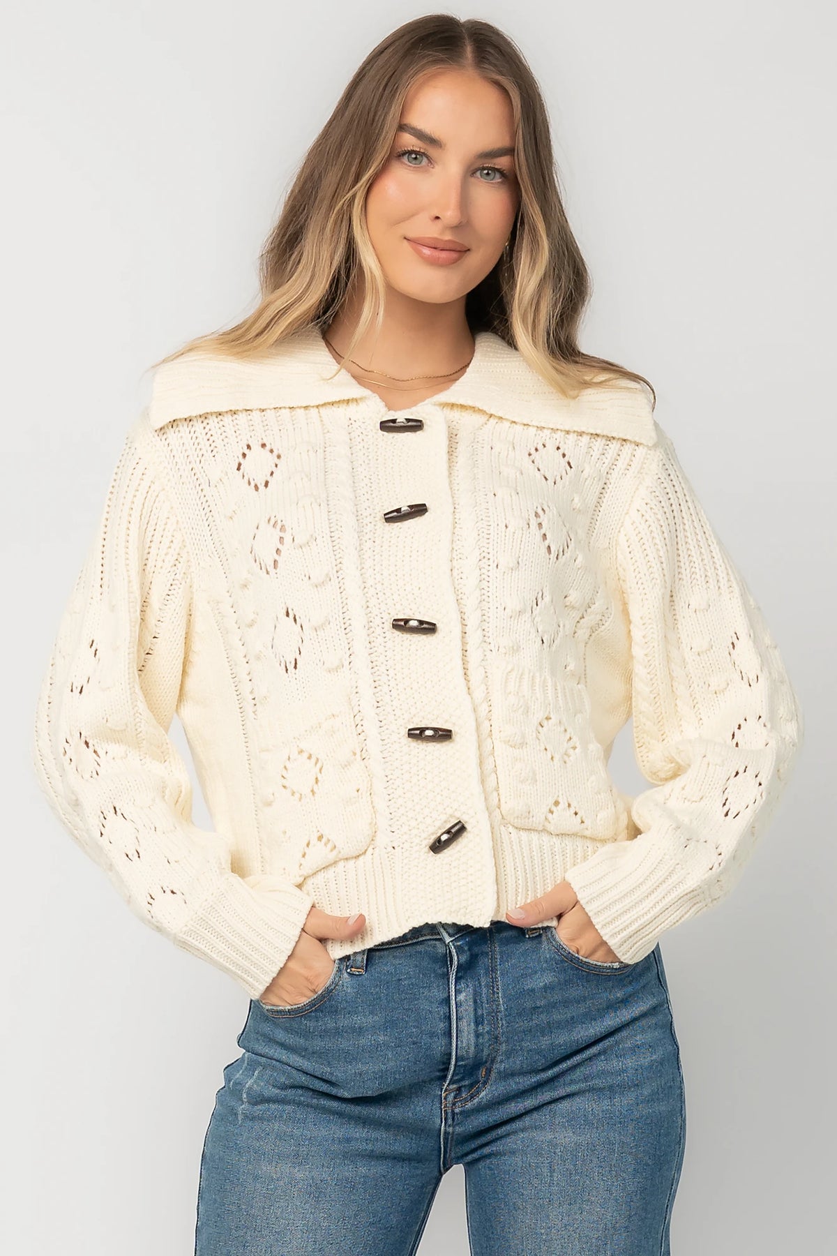 Cream cable knit sweater with a wide fold-over collar, toggle button front, long sleeves, bobble and pointelle knit detailing, ribbed trim, and a relaxed fit.