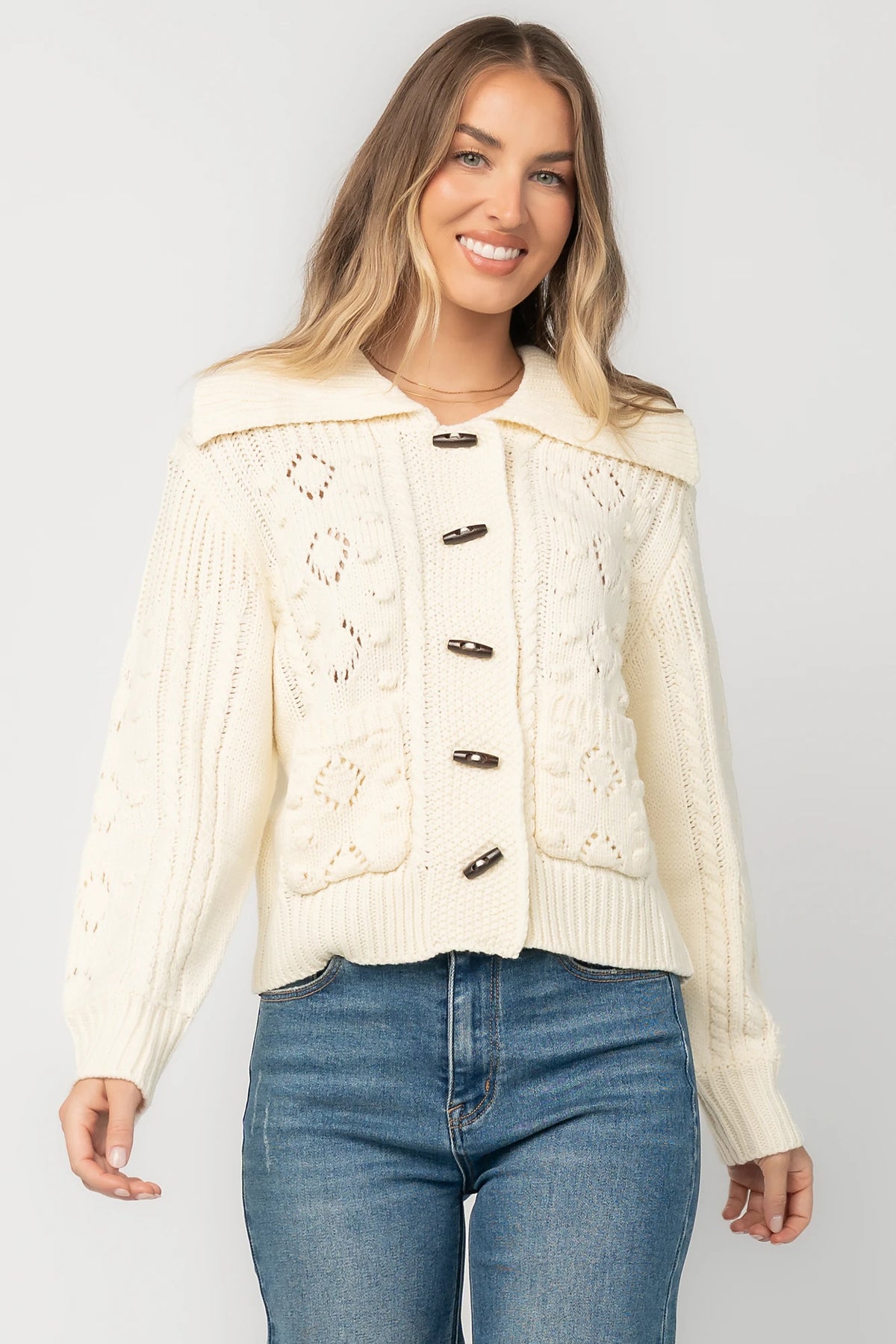 Cream cable knit sweater with a wide fold-over collar, toggle button front, long sleeves, bobble and pointelle knit detailing, ribbed trim, and a relaxed fit.
