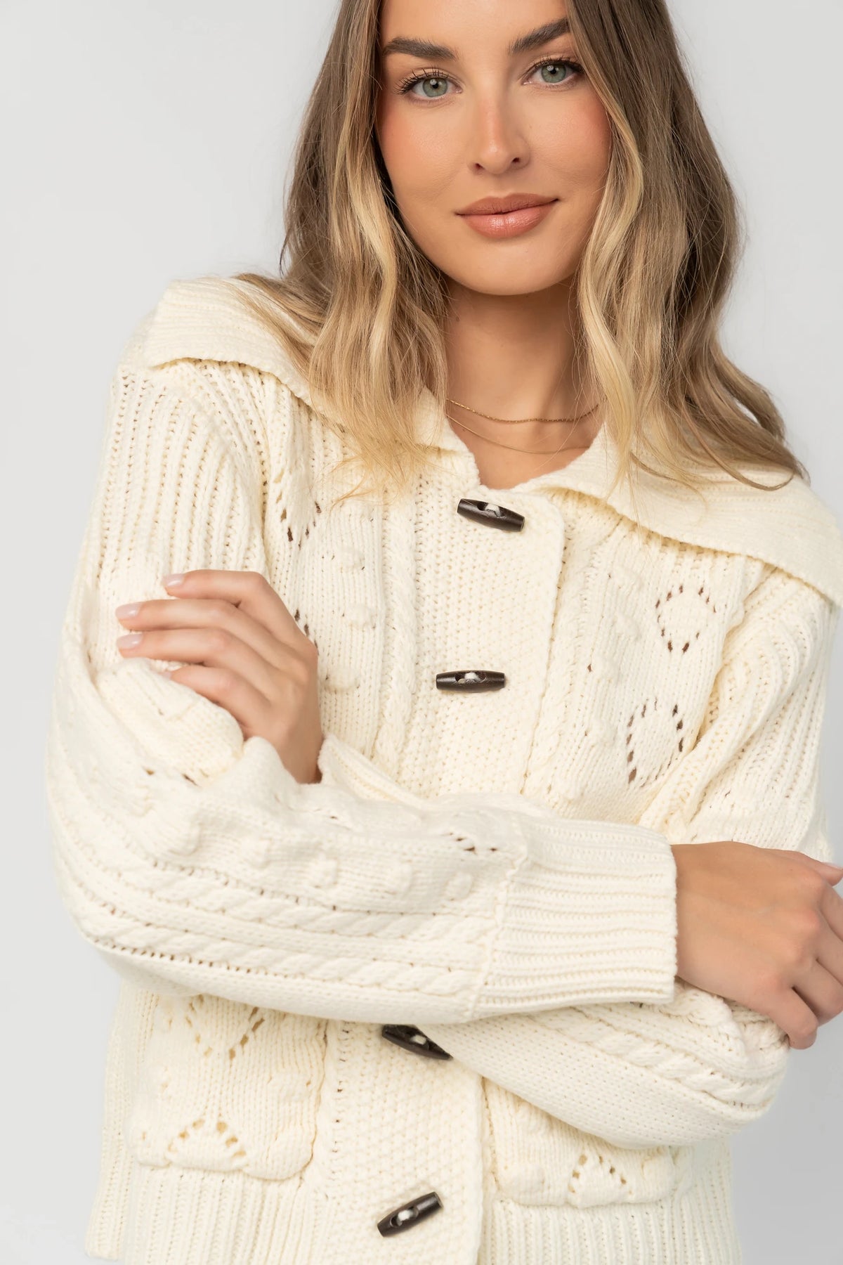 Cream cable knit sweater with a wide fold-over collar, toggle button front, long sleeves, bobble and pointelle knit detailing, ribbed trim, and a relaxed fit.