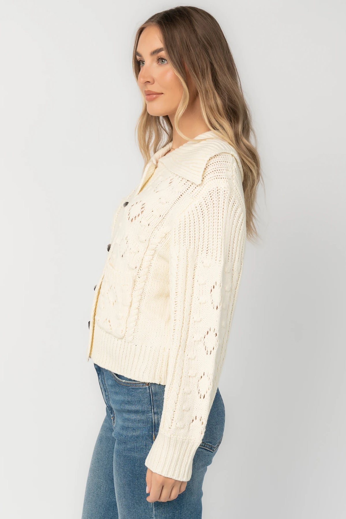Cream cable knit sweater with a wide fold-over collar, toggle button front, long sleeves, bobble and pointelle knit detailing, ribbed trim, and a relaxed fit.