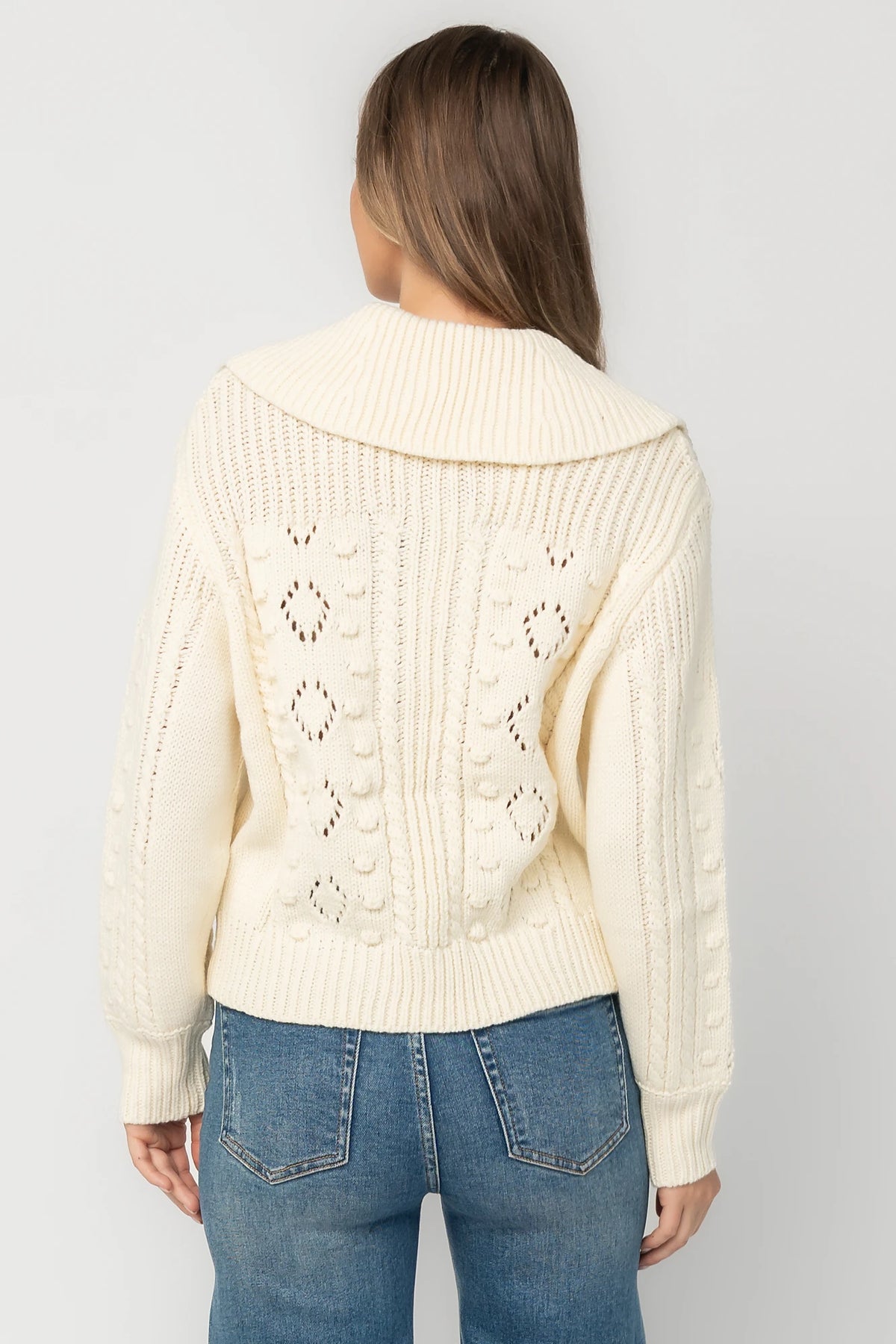 Cream cable knit sweater with a wide fold-over collar, toggle button front, long sleeves, bobble and pointelle knit detailing, ribbed trim, and a relaxed fit.