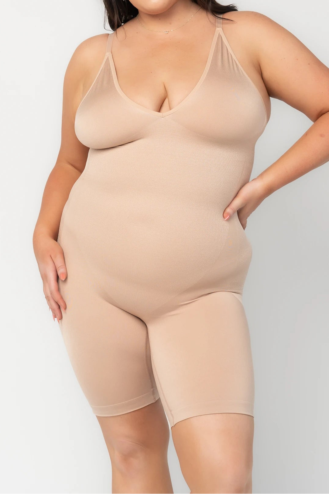 Model wearing a nude shaping bodysuit with adjustable spaghetti straps, a deep V-neckline, and mid-thigh shorts. The seamless design features targeted compression through the waist and abdomen for a smooth, sculpted silhouette. One hand rests at the waist.