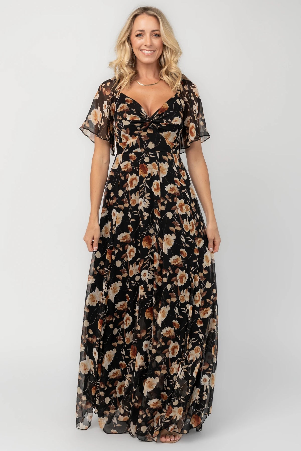 Black floral chiffon maxi dress with a sweetheart neckline, flutter sleeves, smocked back bodice, tiered skirt, and subtle gold foil detailing.
