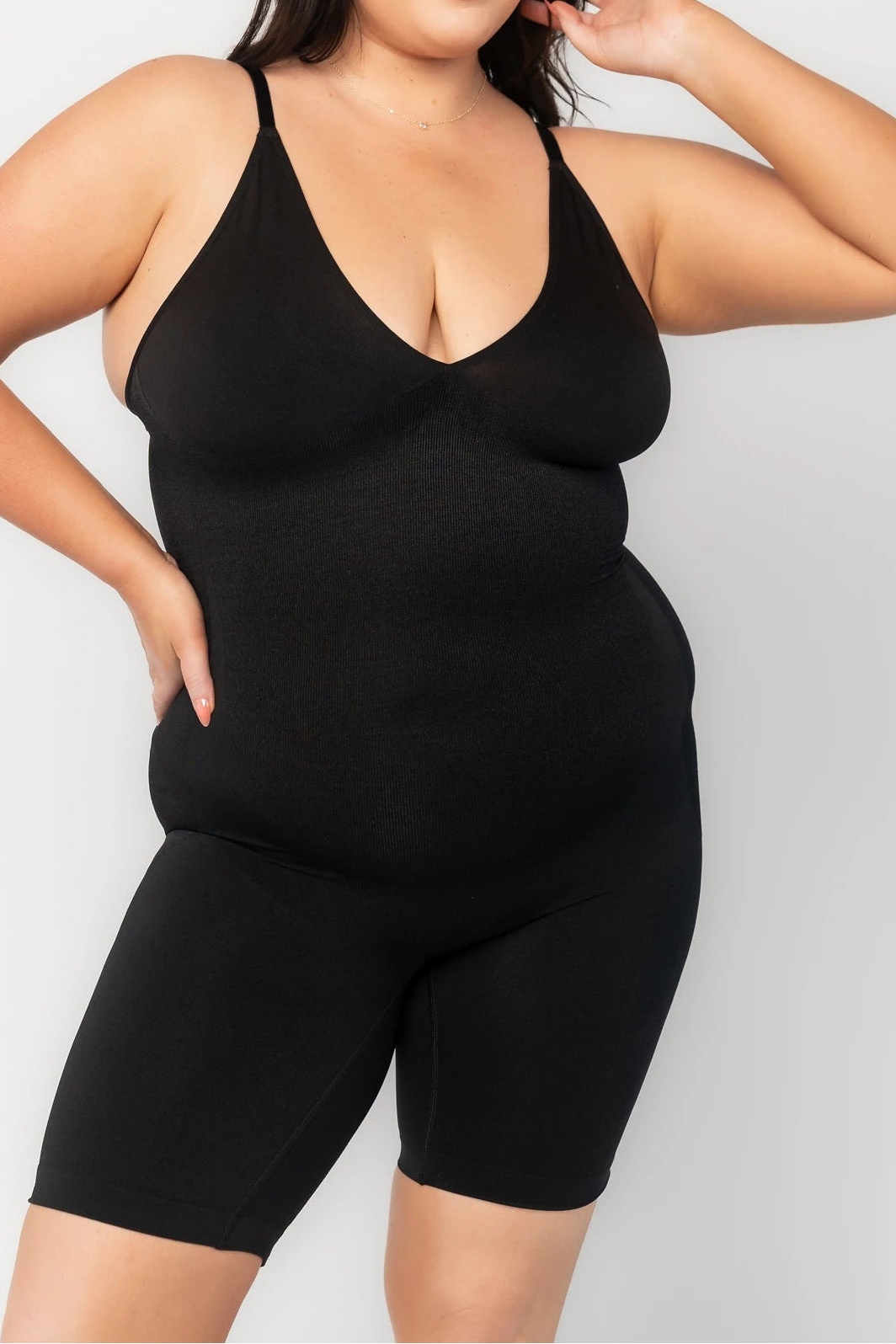 Model wearing a black shaping bodysuit with adjustable spaghetti straps, a deep V-neckline, and mid-thigh shorts. The seamless design features targeted compression through the waist and abdomen for a smooth, sculpted silhouette. 