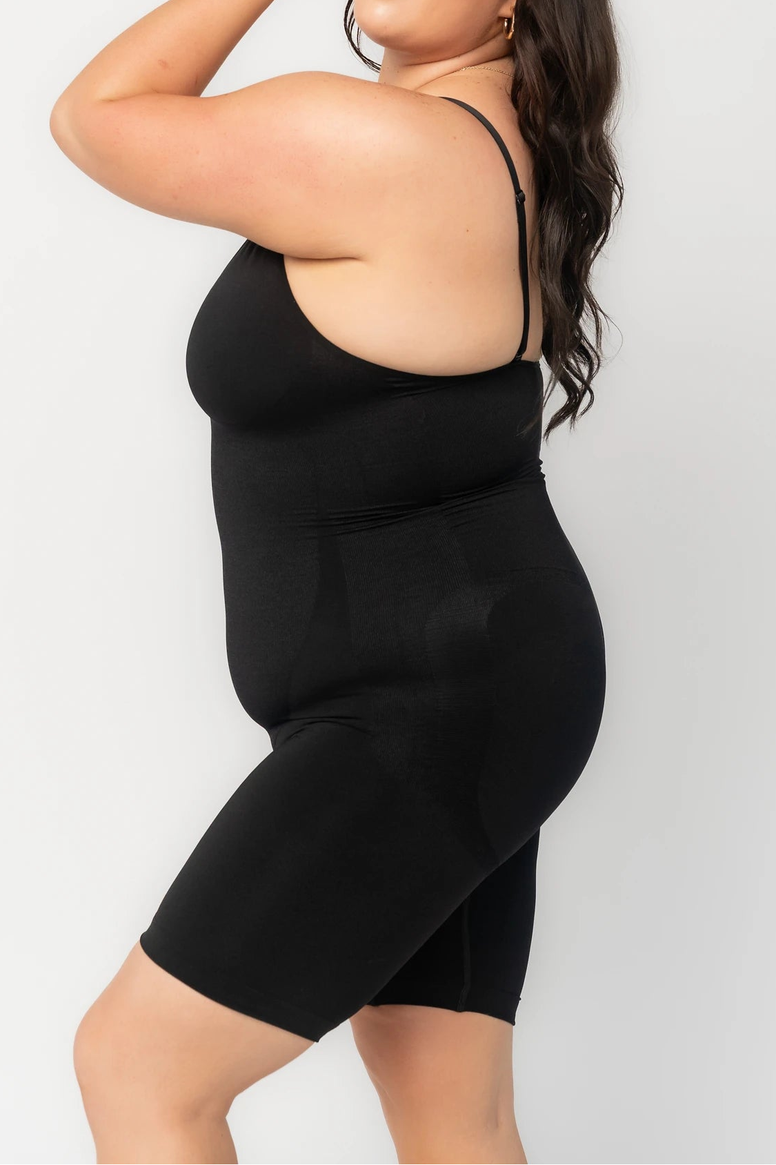 Model wearing a black shaping bodysuit with adjustable spaghetti straps, a deep V-neckline, and mid-thigh shorts. The seamless design features targeted compression through the waist and abdomen for a smooth, sculpted silhouette. 