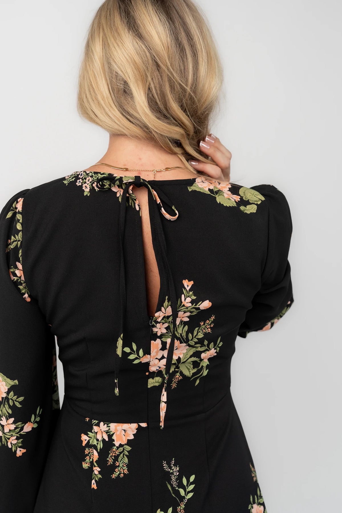 Black floral mini dress with a round neckline, long sleeves, fitted bodice, ruffle hem, back keyhole tie, and hidden back zipper.