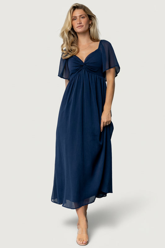 Front view of model wearing navy Nora Midi Dress with sweetheart neckline, knotted bust, empire waist and flutter sleeves, worn with neutral shoes.
