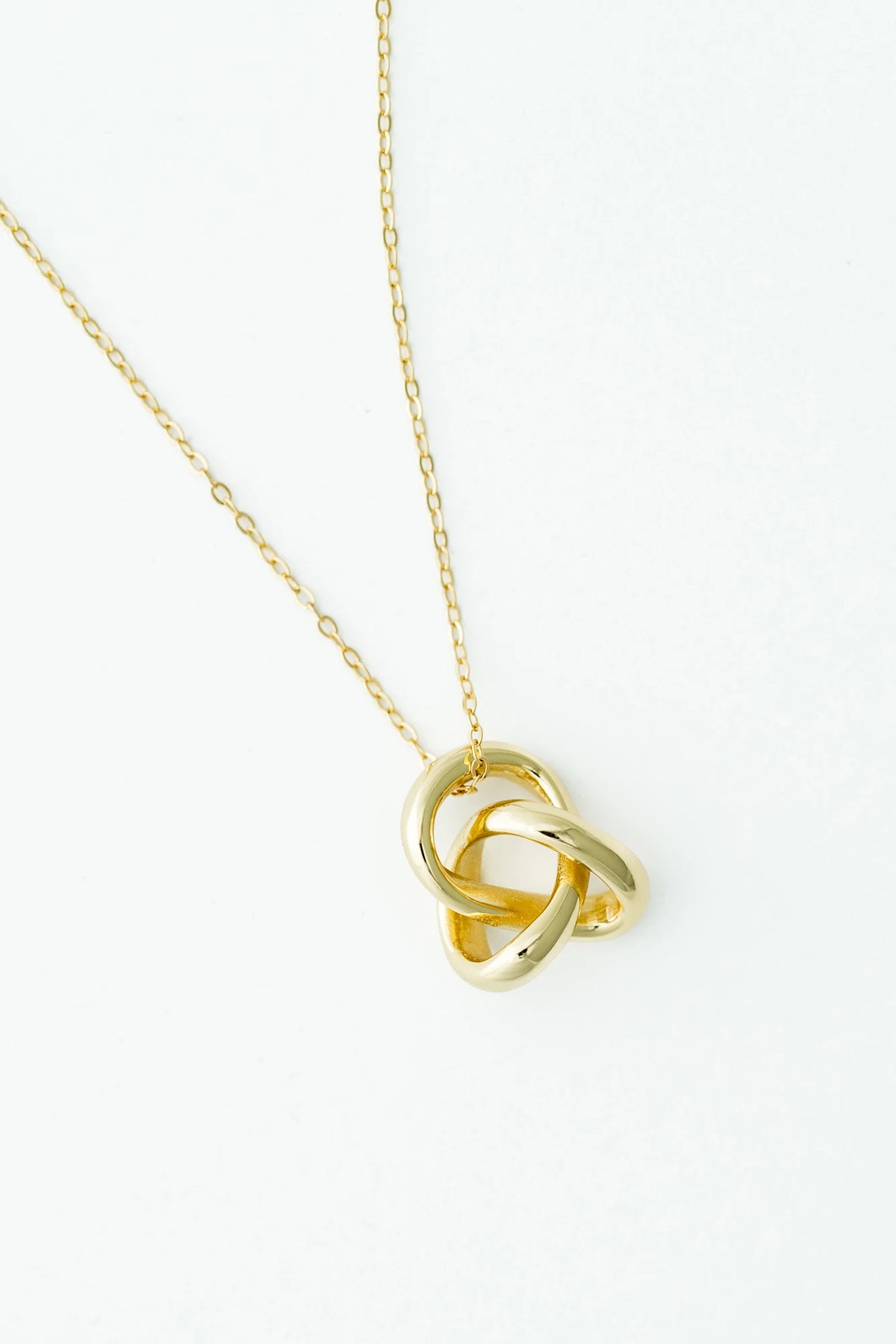 White background of a delicate gold-tone necklace featuring an interlocking infinity knot pendant suspended from a fine chain. 