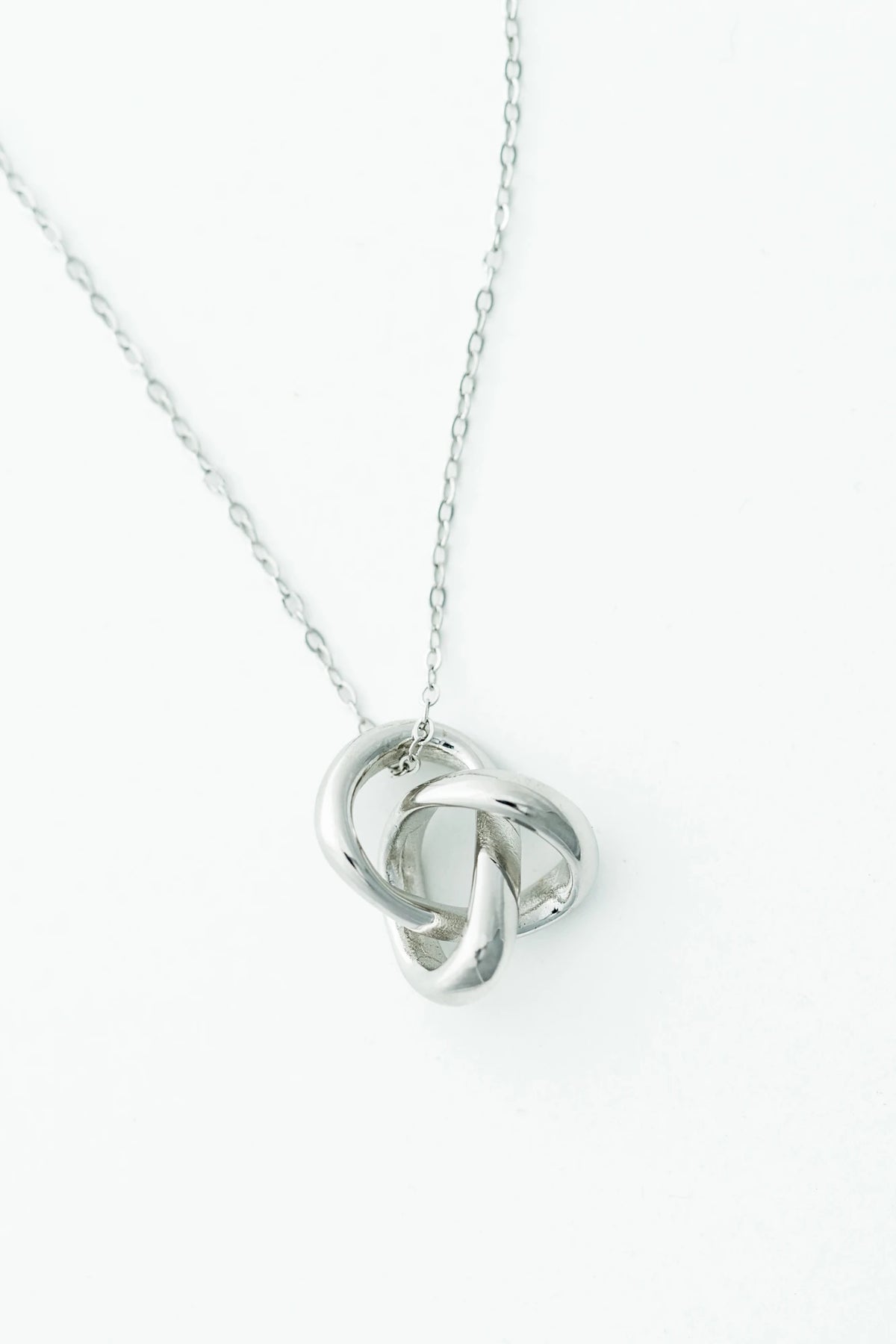 White background of  a delicate silver-tone necklace featuring an interlocking infinity knot pendant suspended from a fine chain. 