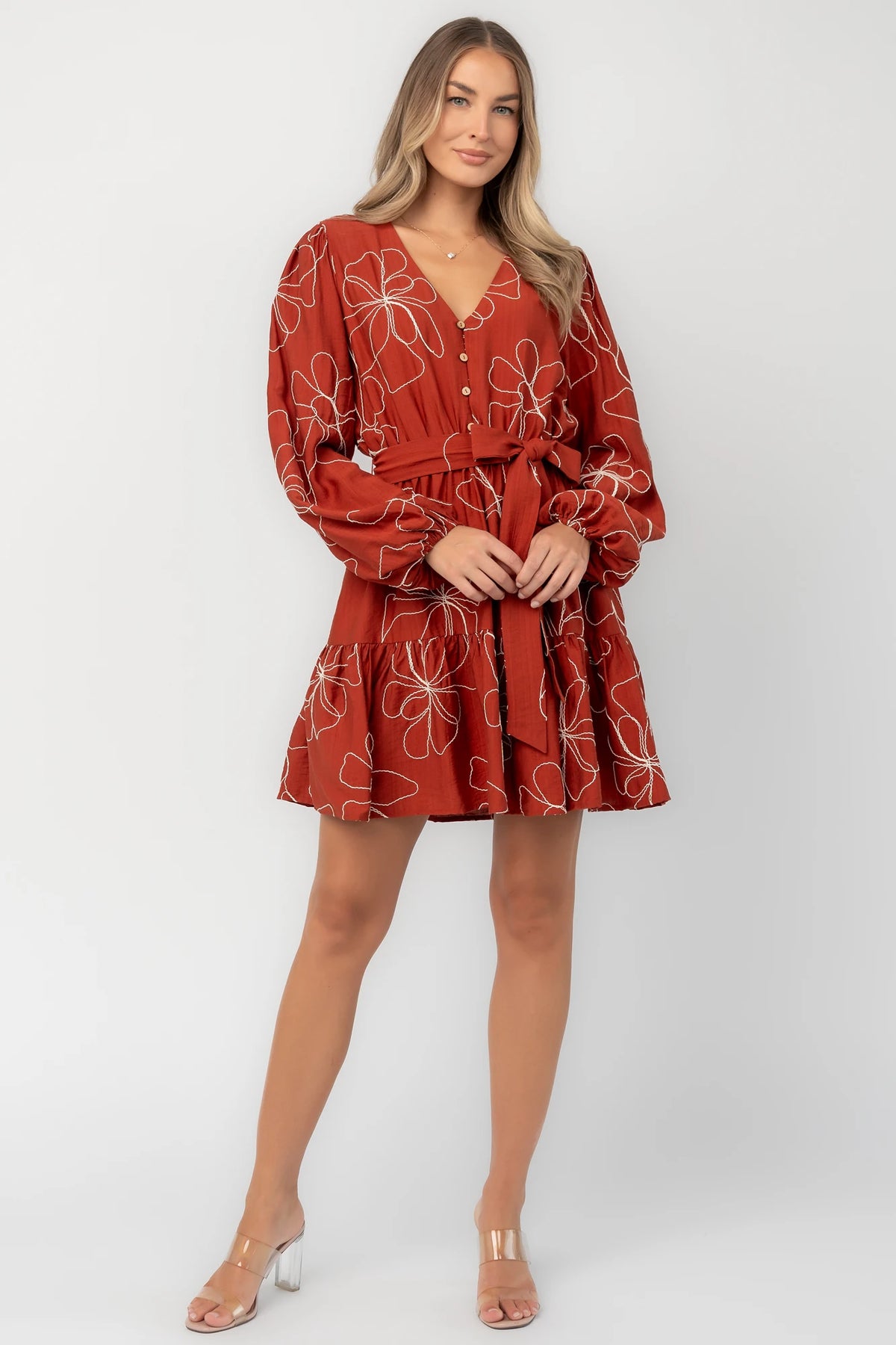 Rust floral mini dress with a V-neckline, button-front bodice, long balloon sleeves, elastic waistband with a self-tie sash, tiered ruffle hem, and side pockets.