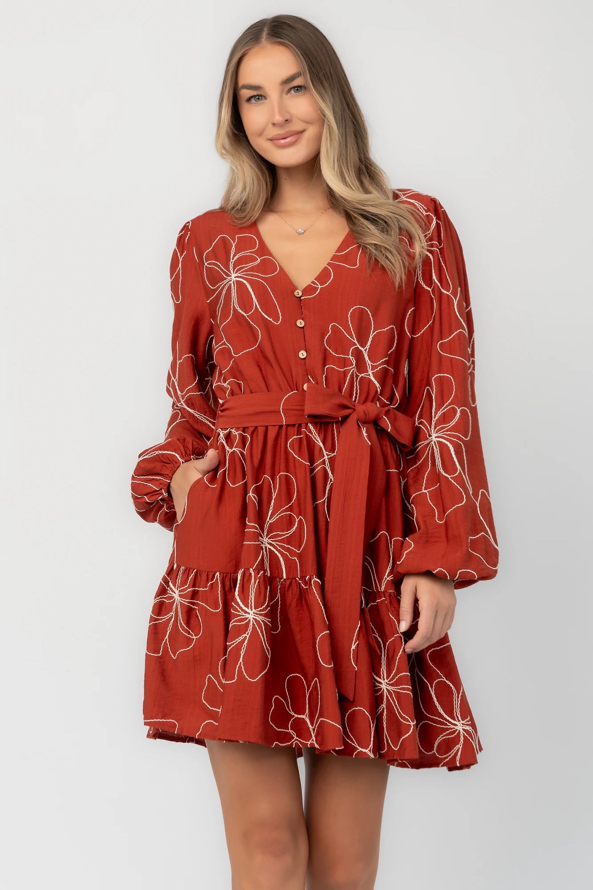 Rust floral mini dress with a V-neckline, button-front bodice, long balloon sleeves, elastic waistband with a self-tie sash, tiered ruffle hem, and side pockets.