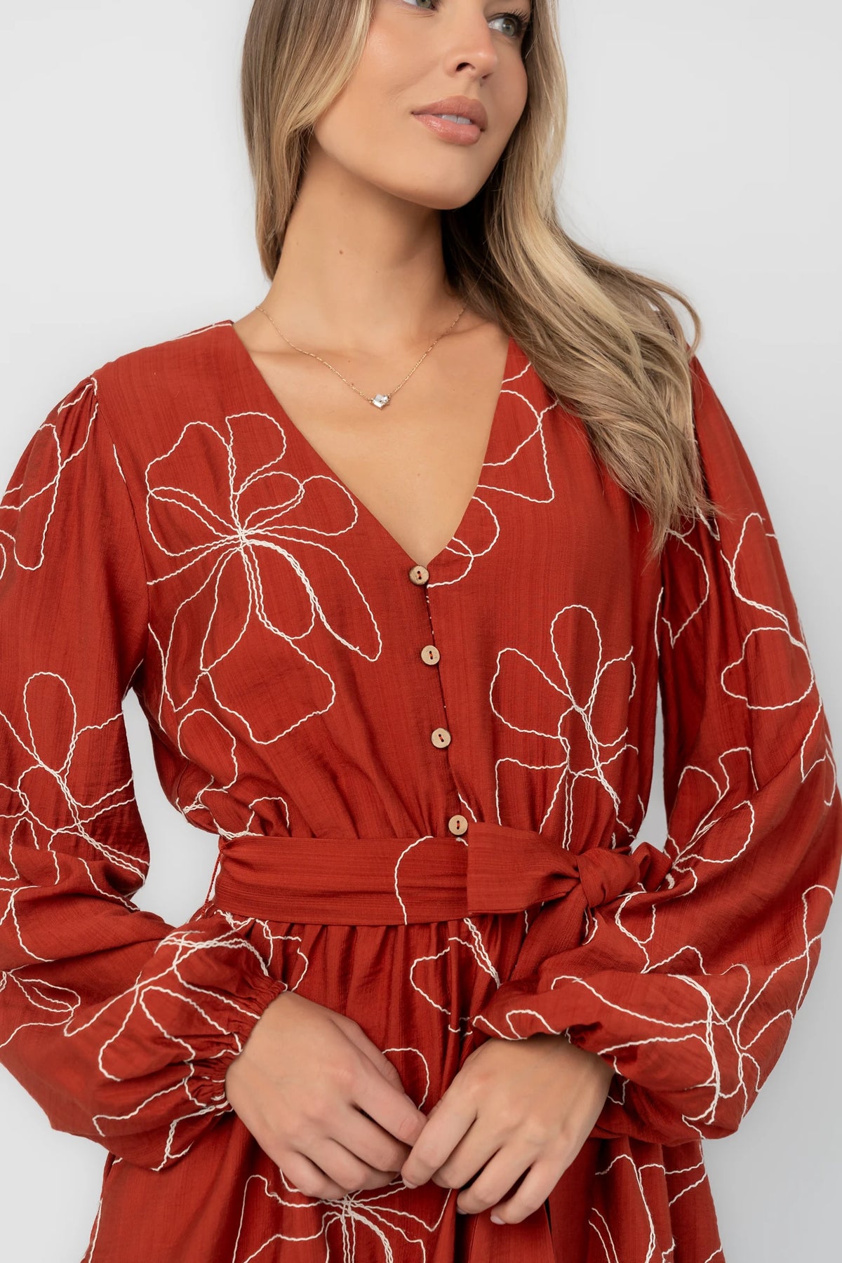 Rust floral mini dress with a V-neckline, button-front bodice, long balloon sleeves, elastic waistband with a self-tie sash, tiered ruffle hem, and side pockets.
