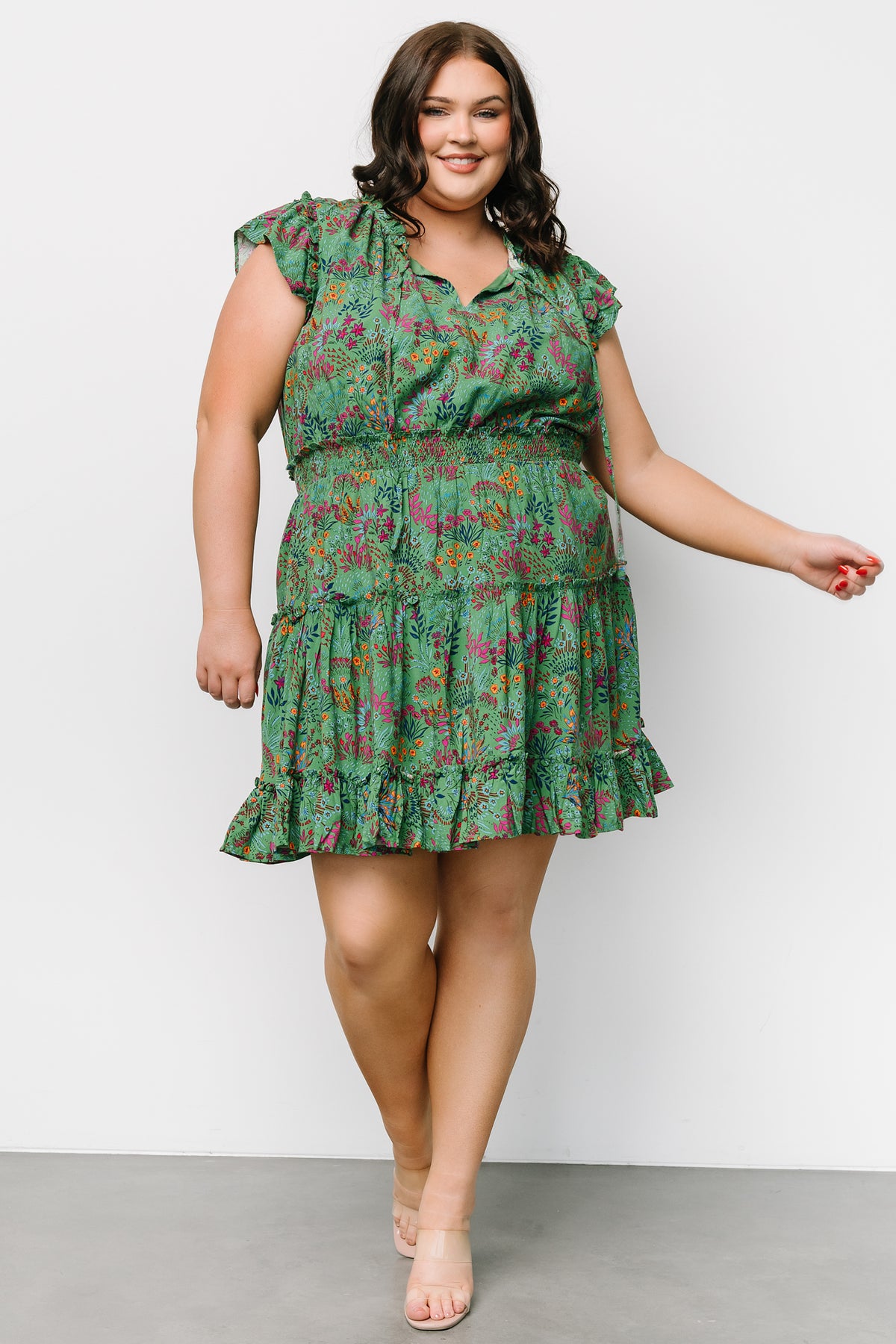 Three-quarter angle of the Rio Short Dress in green multi print, highlighting layered ruffle sleeves, ruffle trim at the top hem, and flowing rayon fabric with lining.