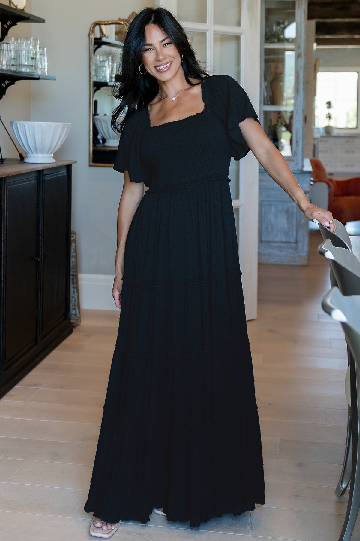 Front view of Lenora smocked dotted maxi dress in black on a model, showing square neckline, smocked bodice and flowing ruffle tiered skirt for elegant movement.