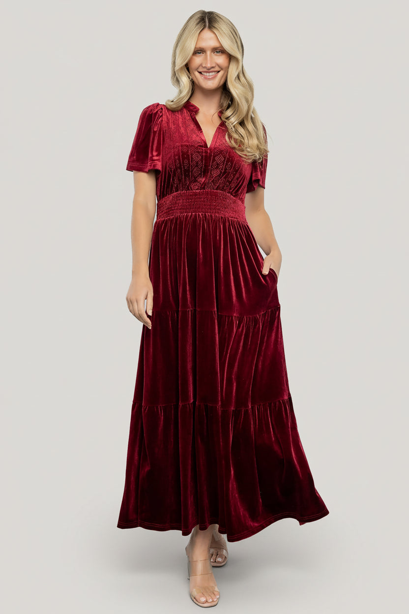 Front model view of Maida cranberry velvet maxi dress showing V-neck collar, embroidered front bodice, smocked waistband and flowing tiered skirt for movement.