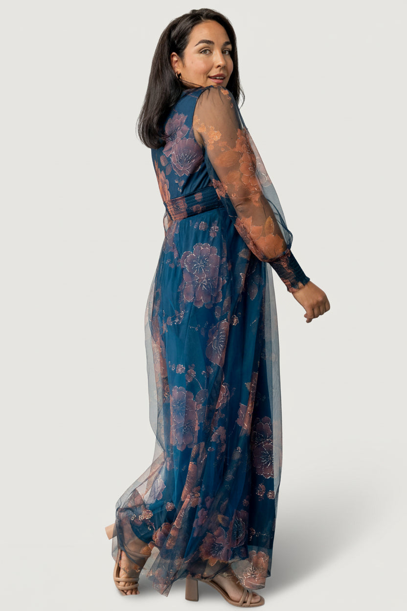 Model seated wearing copper and blue metallic tulle maxi dress, demonstrating flexible smocked waist, bump-friendly silhouette, and sleeve sheerness.