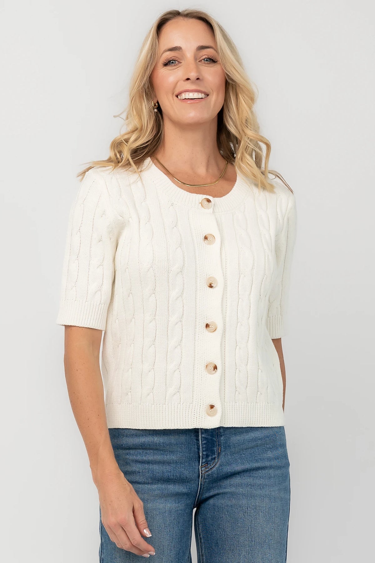 Cream cable knit cardigan with a round neckline, button-front closure, short sleeves, ribbed trim, and a fitted silhouette.