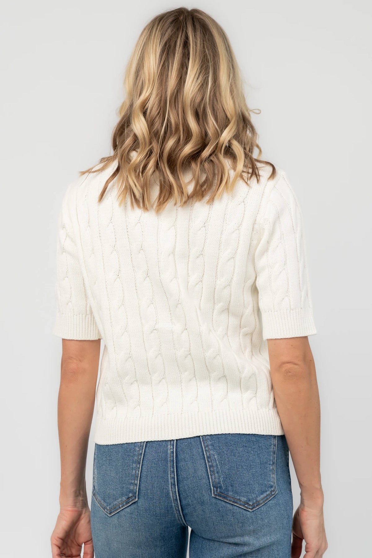 Cream cable knit cardigan with a round neckline, button-front closure, short sleeves, ribbed trim, and a fitted silhouette.