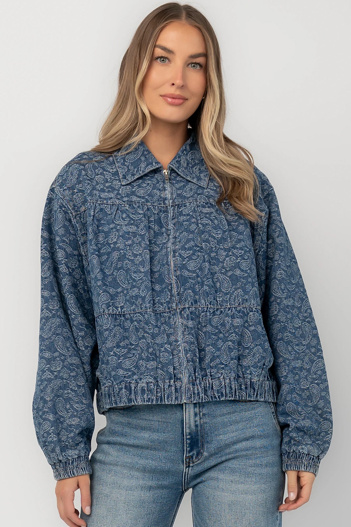Medium wash paisley print denim jacket with a collared neckline, front zipper closure, long sleeves, elastic hem, and side pockets.