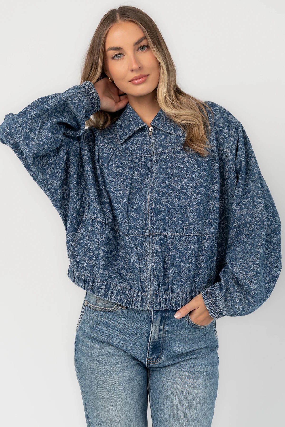 Medium wash paisley print denim jacket with a collared neckline, front zipper closure, long sleeves, elastic hem, and side pockets.
