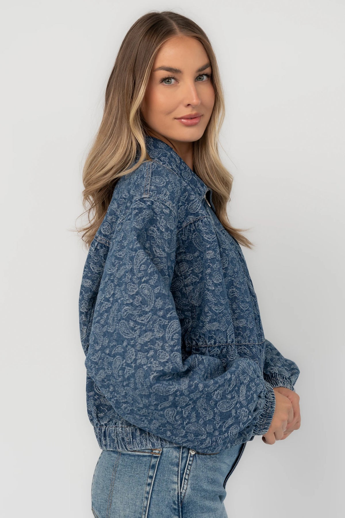 Medium wash paisley print denim jacket with a collared neckline, front zipper closure, long sleeves, elastic hem, and side pockets.