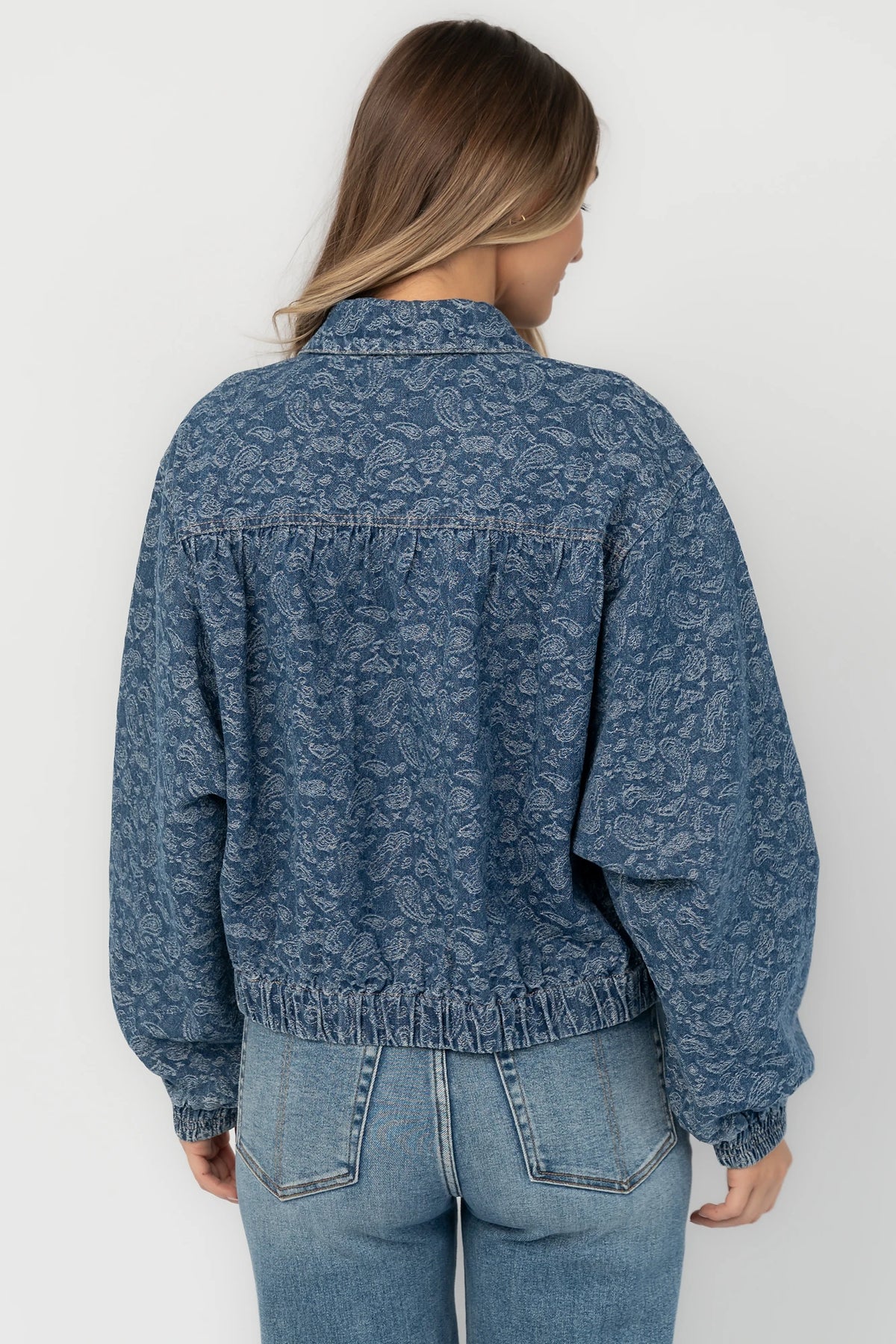 Medium wash paisley print denim jacket with a collared neckline, front zipper closure, long sleeves, elastic hem, and side pockets.