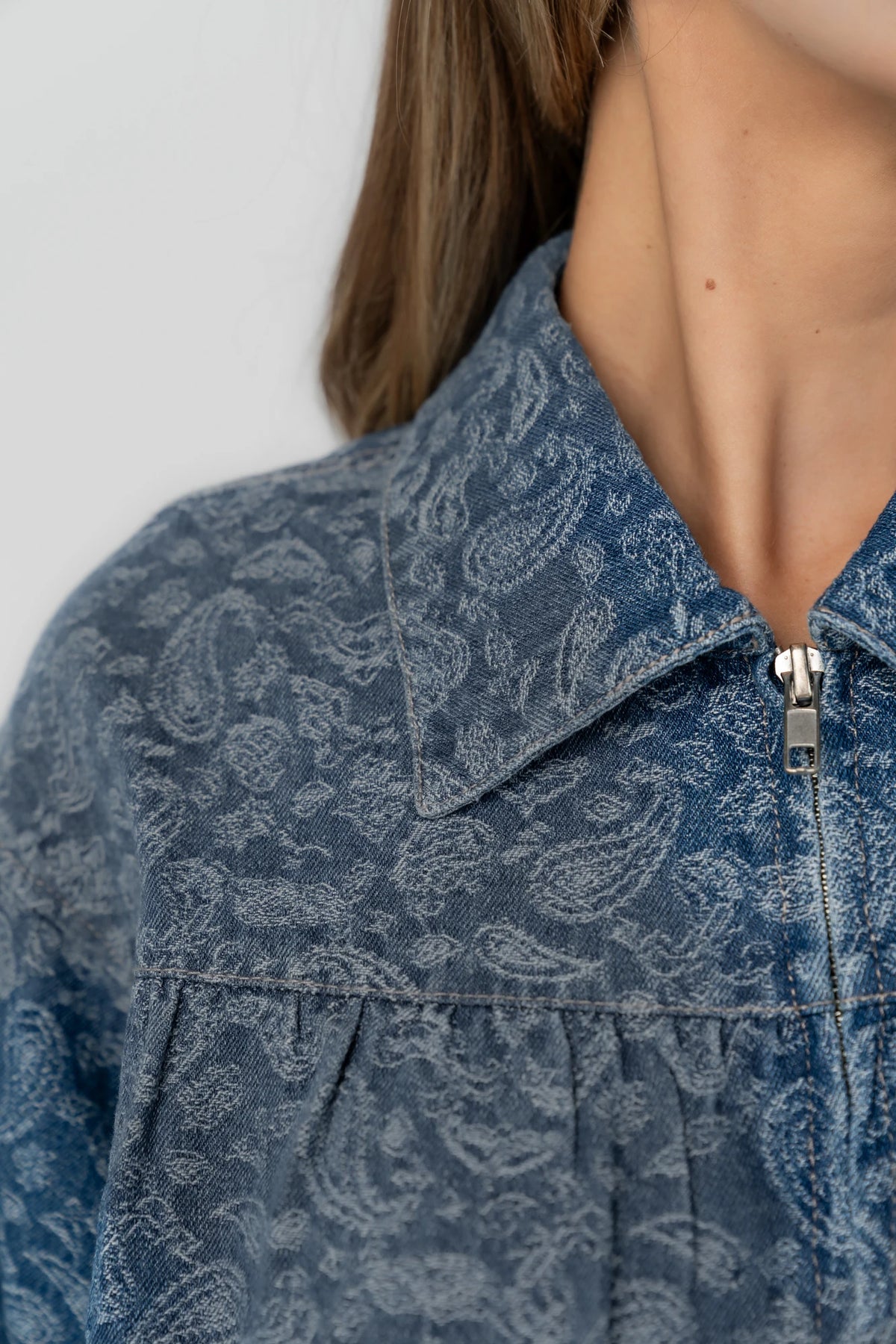 Medium wash paisley print denim jacket with a collared neckline, front zipper closure, long sleeves, elastic hem, and side pockets.