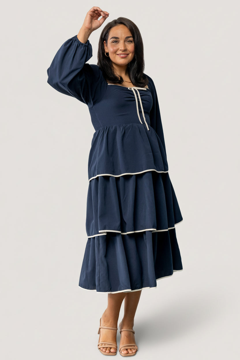 Front view of model wearing navy Barcelona tiered midi dress with sweetheart neckline, decorative bow, and ivory contrast trim at hem and sleeves.