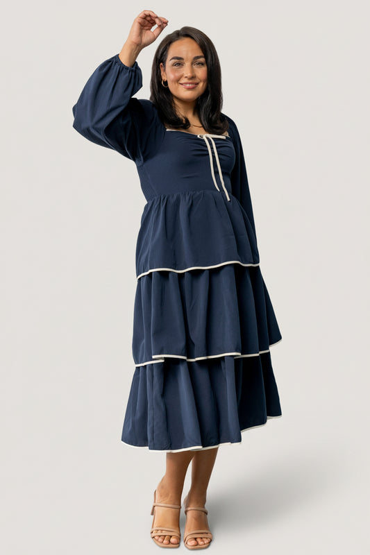 Front view of model wearing navy Barcelona tiered midi dress with sweetheart neckline, decorative bow, and ivory contrast trim at hem and sleeves.
