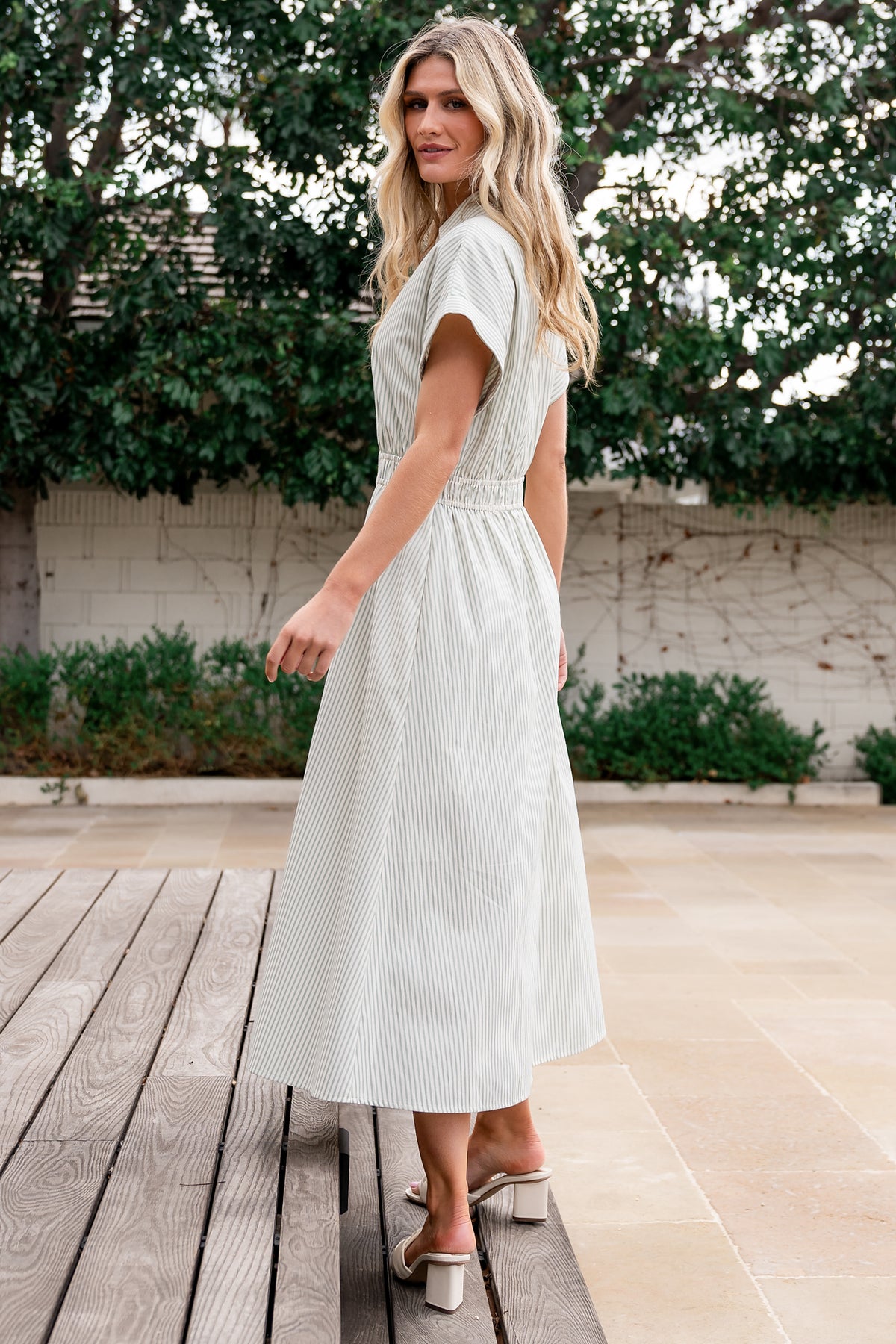 Model walking in sage stripe midi dress with faux-button front and front slit, illustrating movement and midi length.