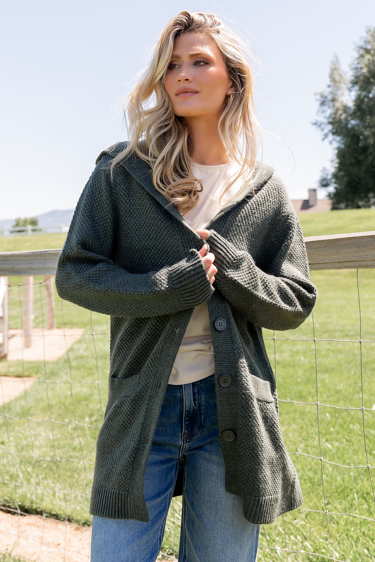 Front view of model wearing deep green Katrine hooded chunky cardigan showing hood, four buttons, and front pockets, styled with jeans.