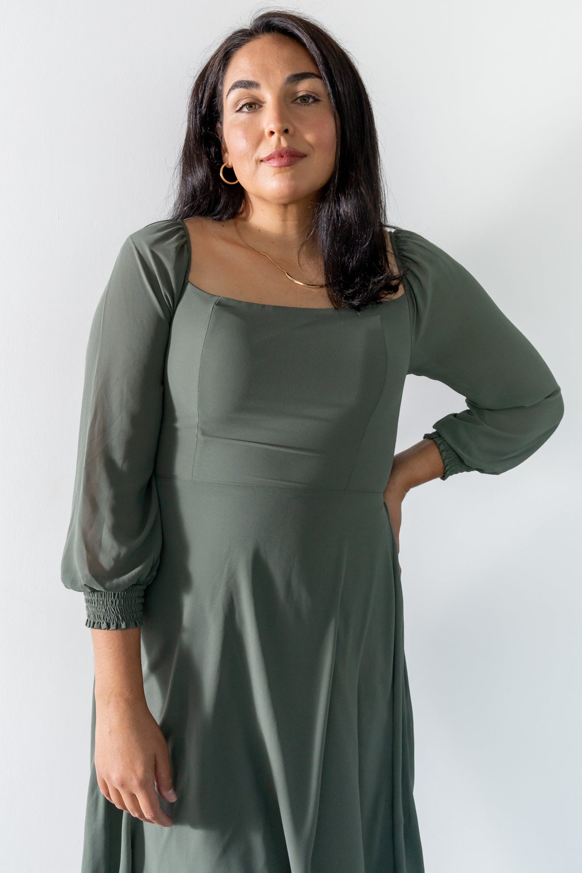 Woman modeling a dark sage chiffon dress with defined waist and flowing skirt, styled with neutral heels