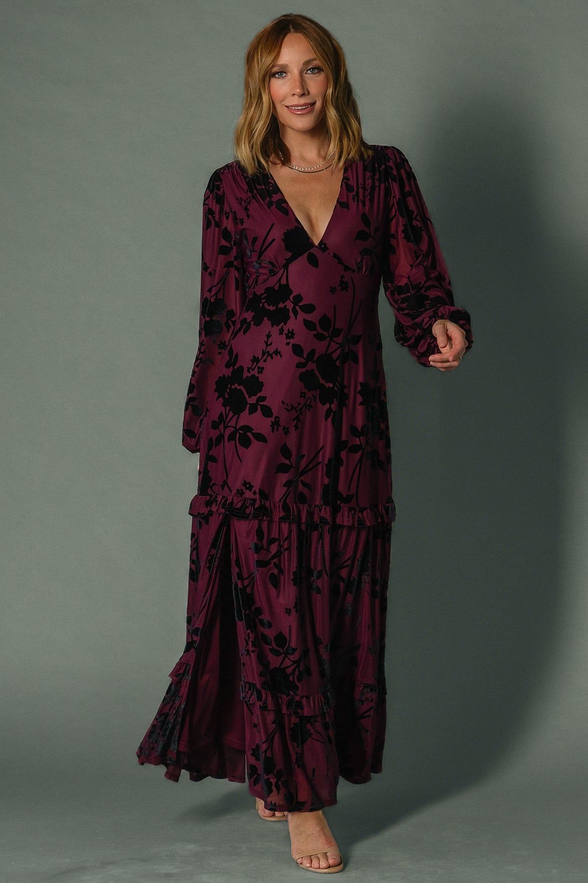 Model wearing mulberry velvet burnout embossed maxi dress, long sleeves, plunging V-neck, ruffle tiered skirt flowing to ankles.