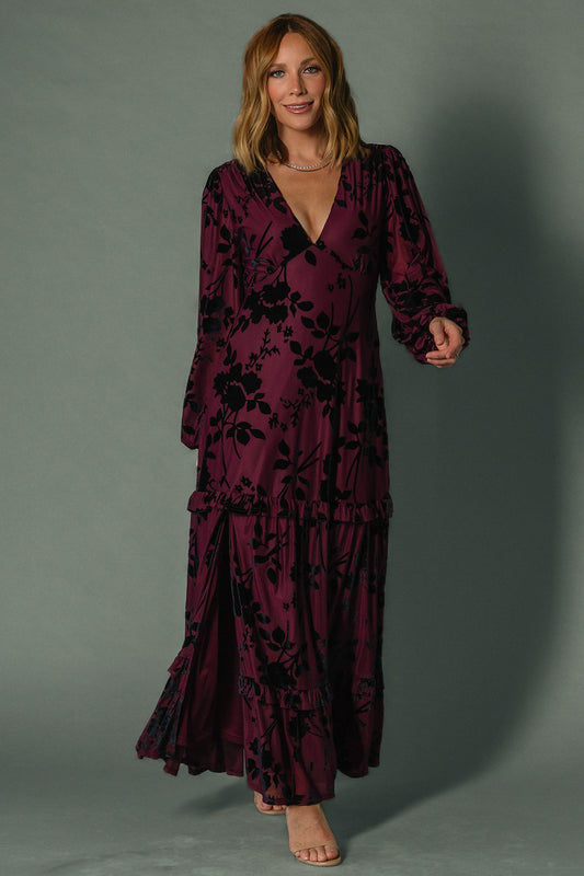 Model wearing mulberry velvet burnout embossed maxi dress, long sleeves, plunging V-neck, ruffle tiered skirt flowing to ankles.