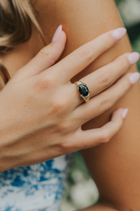 Close-up of Maddie ring on model's hand showing oval black onyx center and gold-toned sculpted wrap-style band, polished finish.