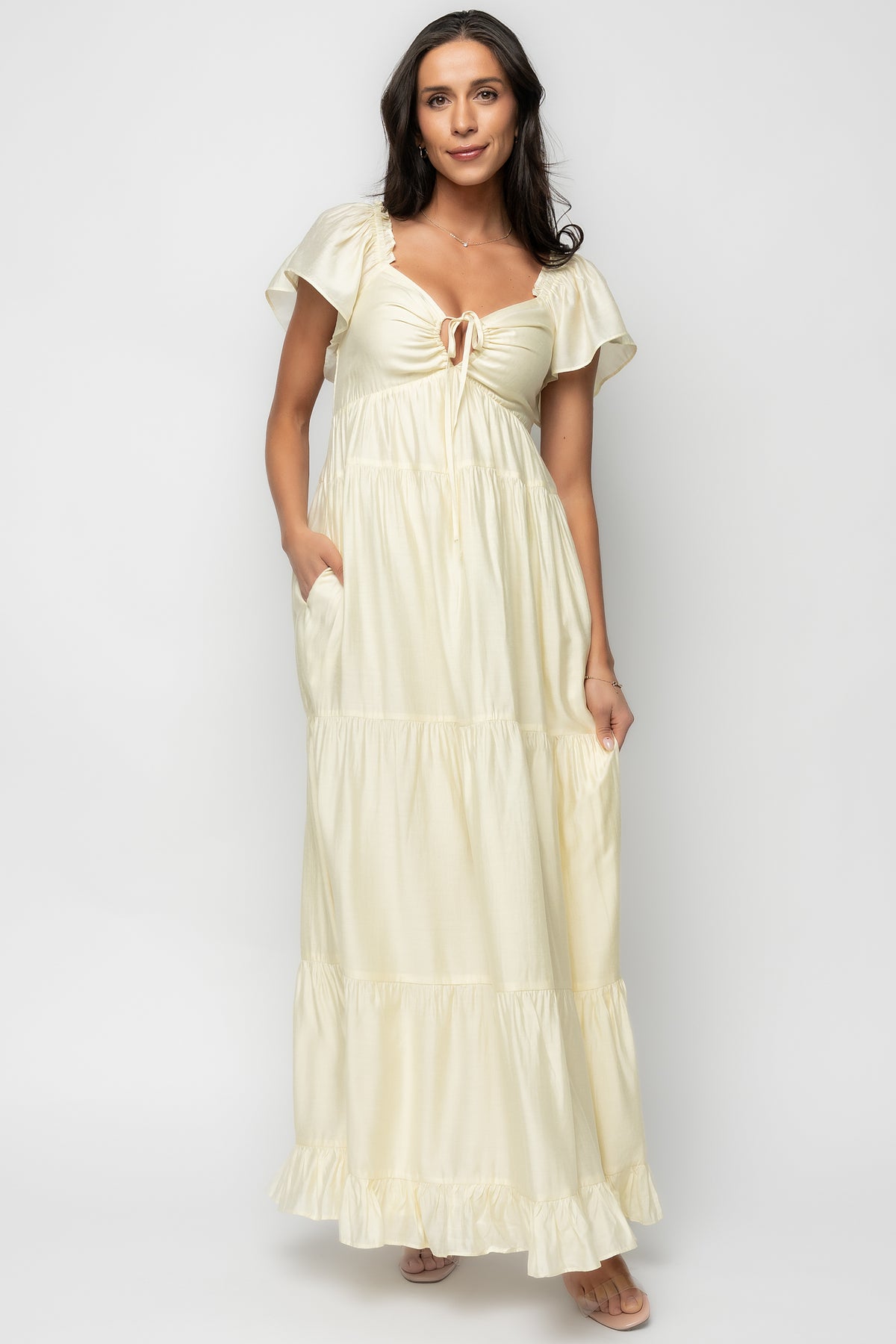 Front full-length view of model wearing pale yellow Agatha Maxi Dress with ruffled V-neck, keyhole tie, short sleeves, and tiered skirt.