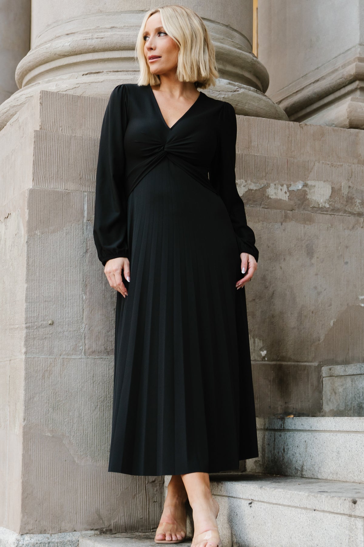 Front view of model wearing Lynda pleated black midi dress with deep V-neck, gathered knot front and long sleeves, shown full length.