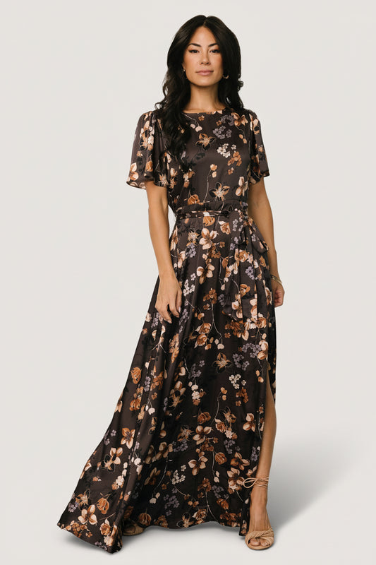 Front view of brown floral satin maxi dress on mannequin featuring rounded neckline, short flutter sleeves, removable belt tie, and left side slit.