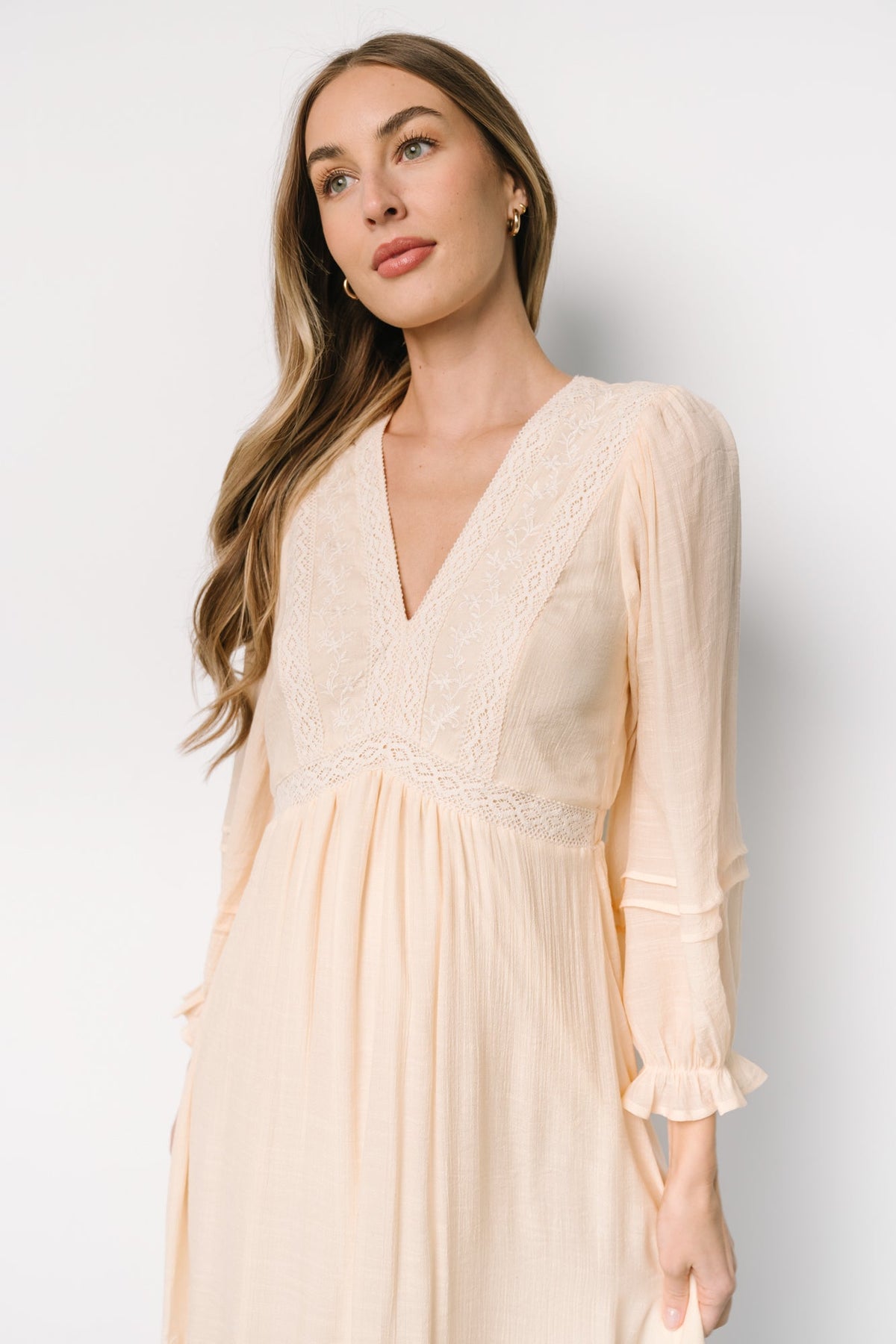 Three-quarter view of Amberlee embroidered cream dress highlighting long ruffled sleeves, V-neckline, and embroidered waist accents.