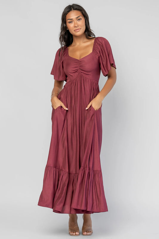Anisa Midi Dress in Burgundy with a wide V-neckline, cinched bust, elastic shoulders, and a tiered skirt.