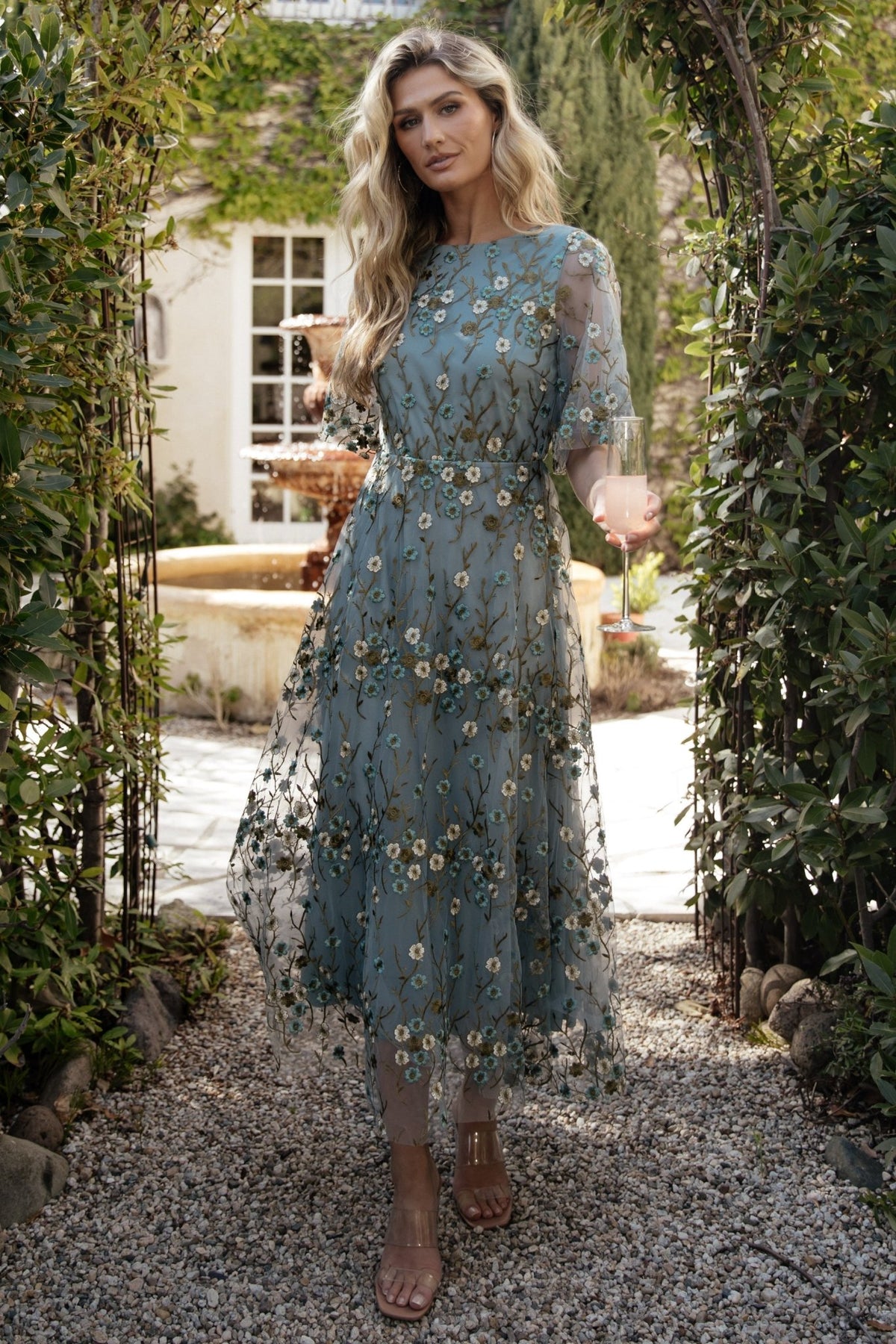 Model wearing Arabella dusty blue floral maxi dress, full-length shot showing flowing skirt, short sheer sleeves, and flattering waist.