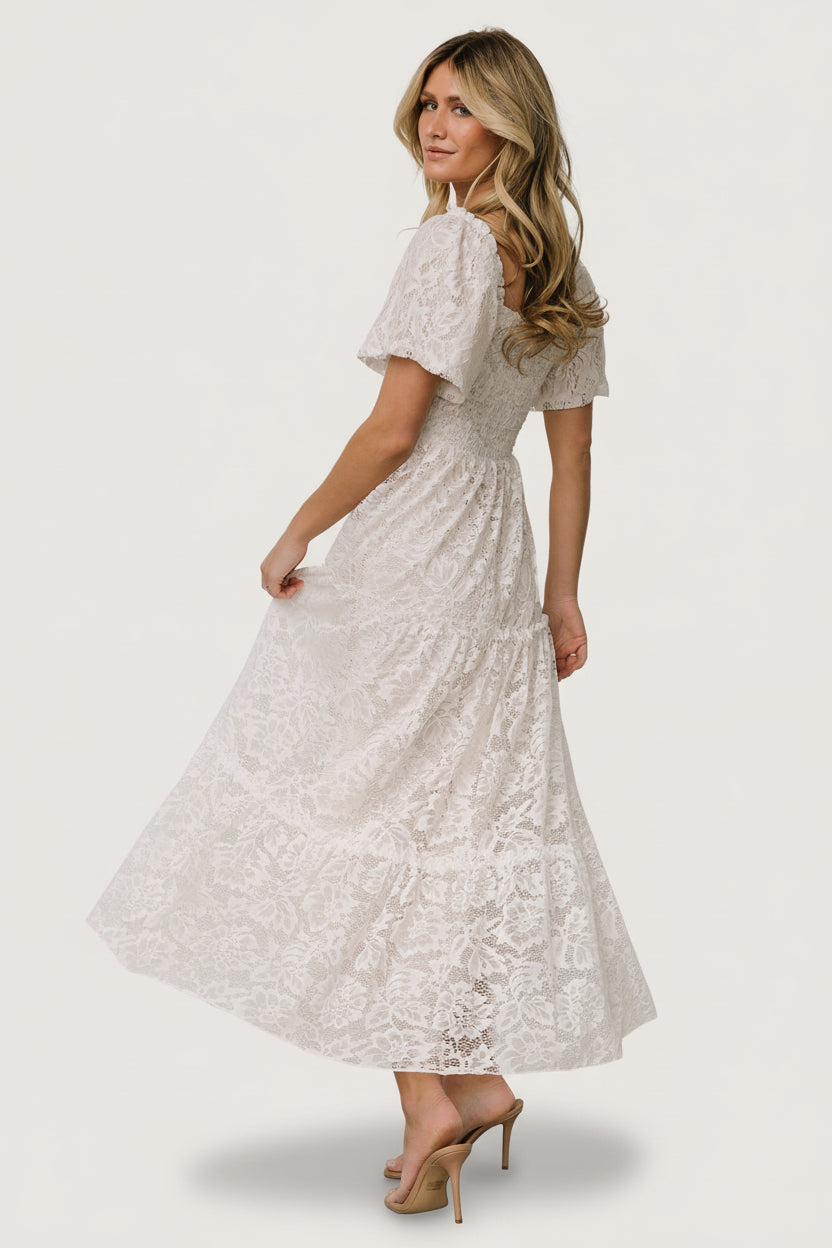 Styled flat-lay of off-white lace maxi dress with visible smocked bodice, ruffle neckline, and layered skirt panels, on a pale surface.