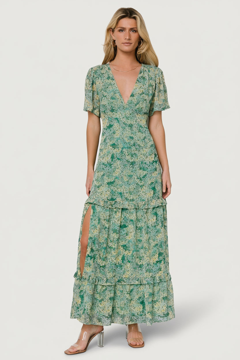Front view of model wearing green floral chiffon deep V maxi dress with tiered ruffle skirt and right-side slit, shown full length.