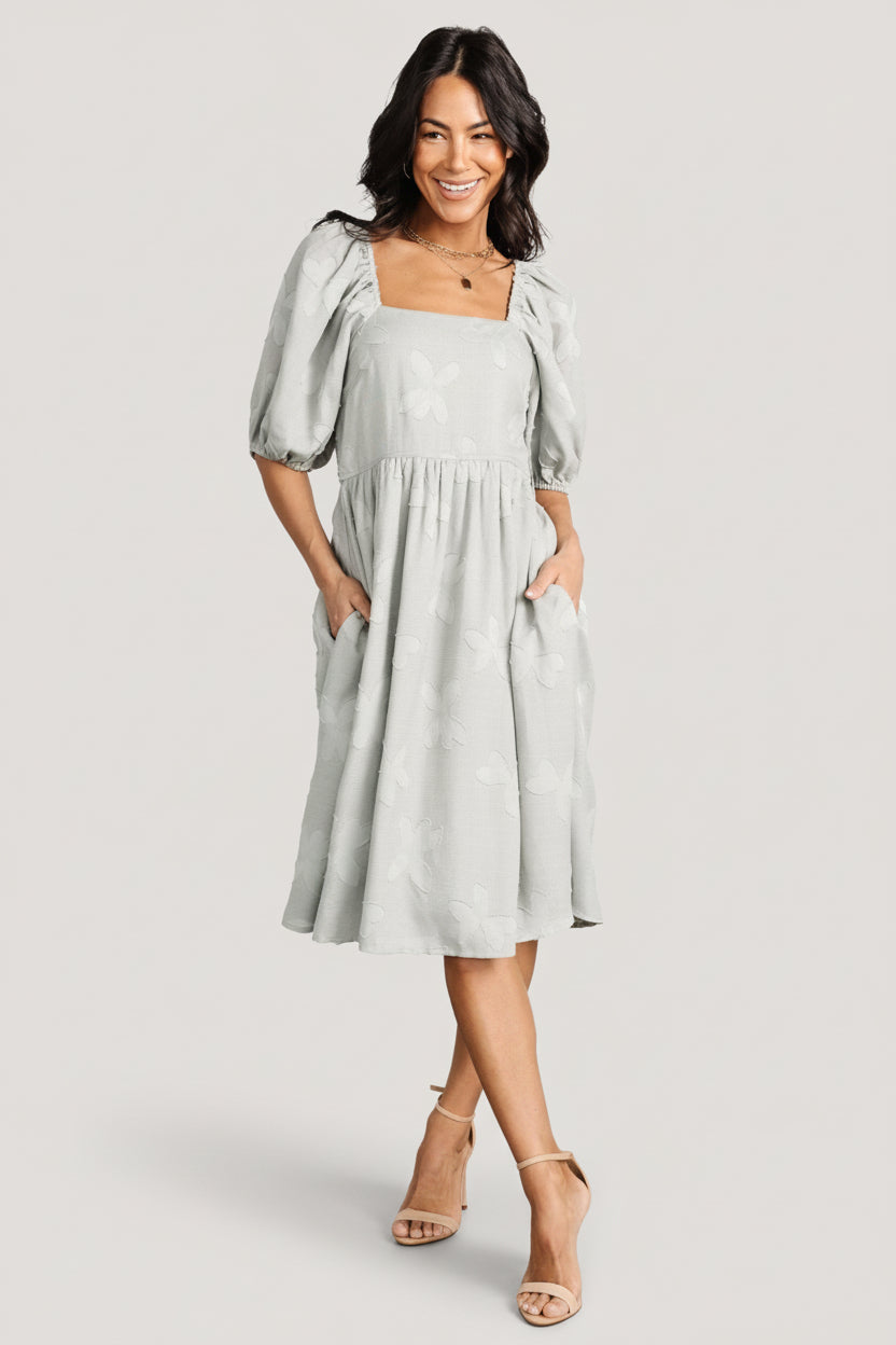 Front view of sage floral short dress with square neckline, smocked back, mid-length puff sleeves, and side pockets.