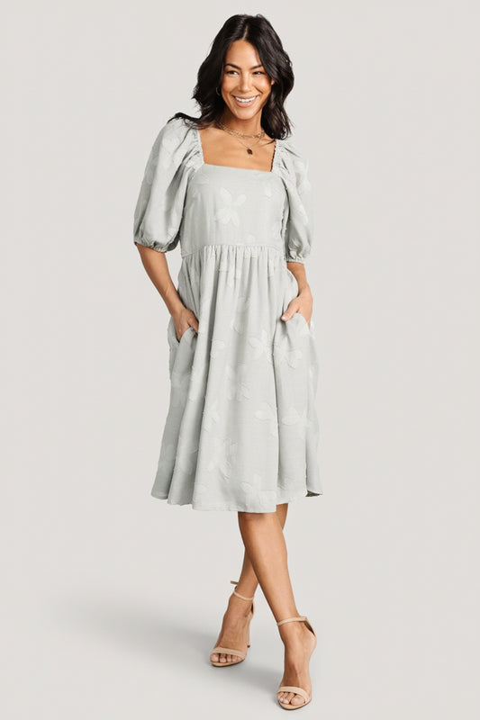 Front view of sage floral short dress with square neckline, smocked back, mid-length puff sleeves, and side pockets.