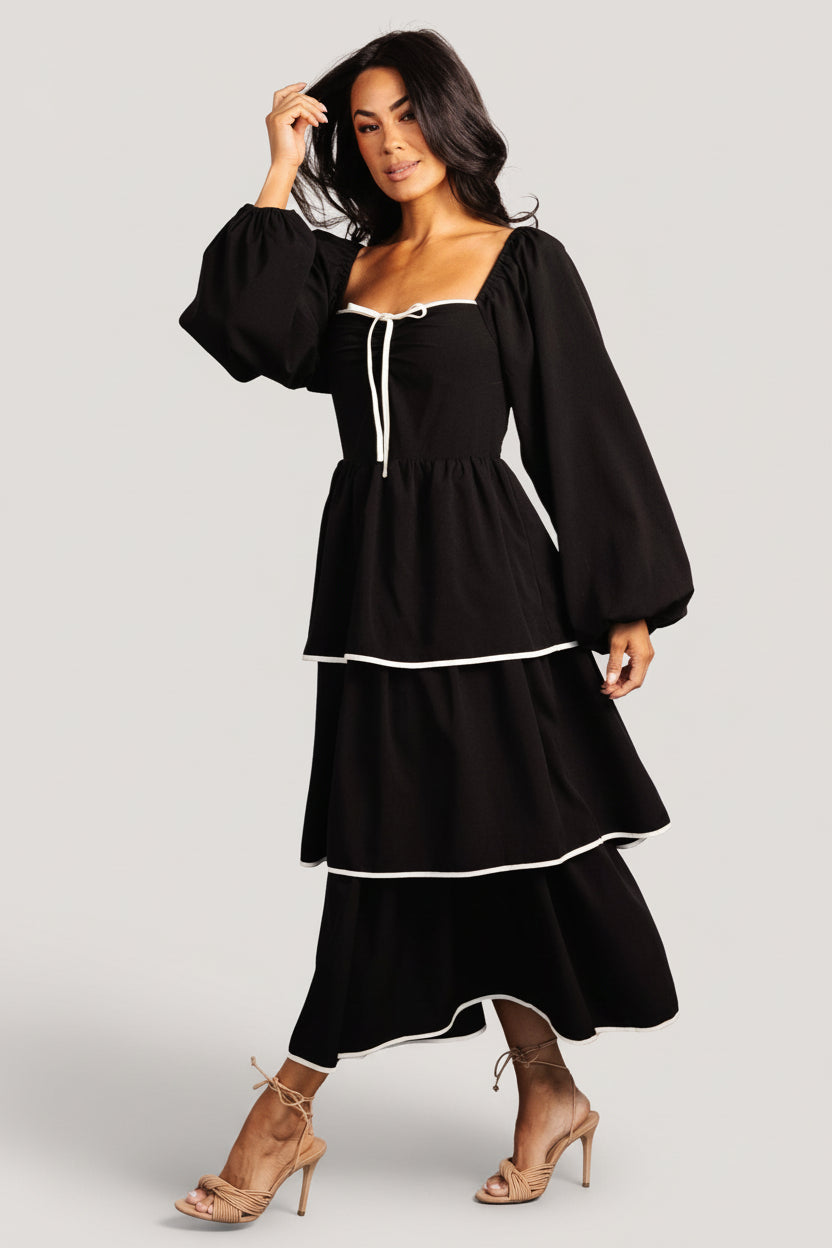 Front view of black tiered midi dress on model, showing soft sweetheart neckline and decorative bow at center bust, full-length.