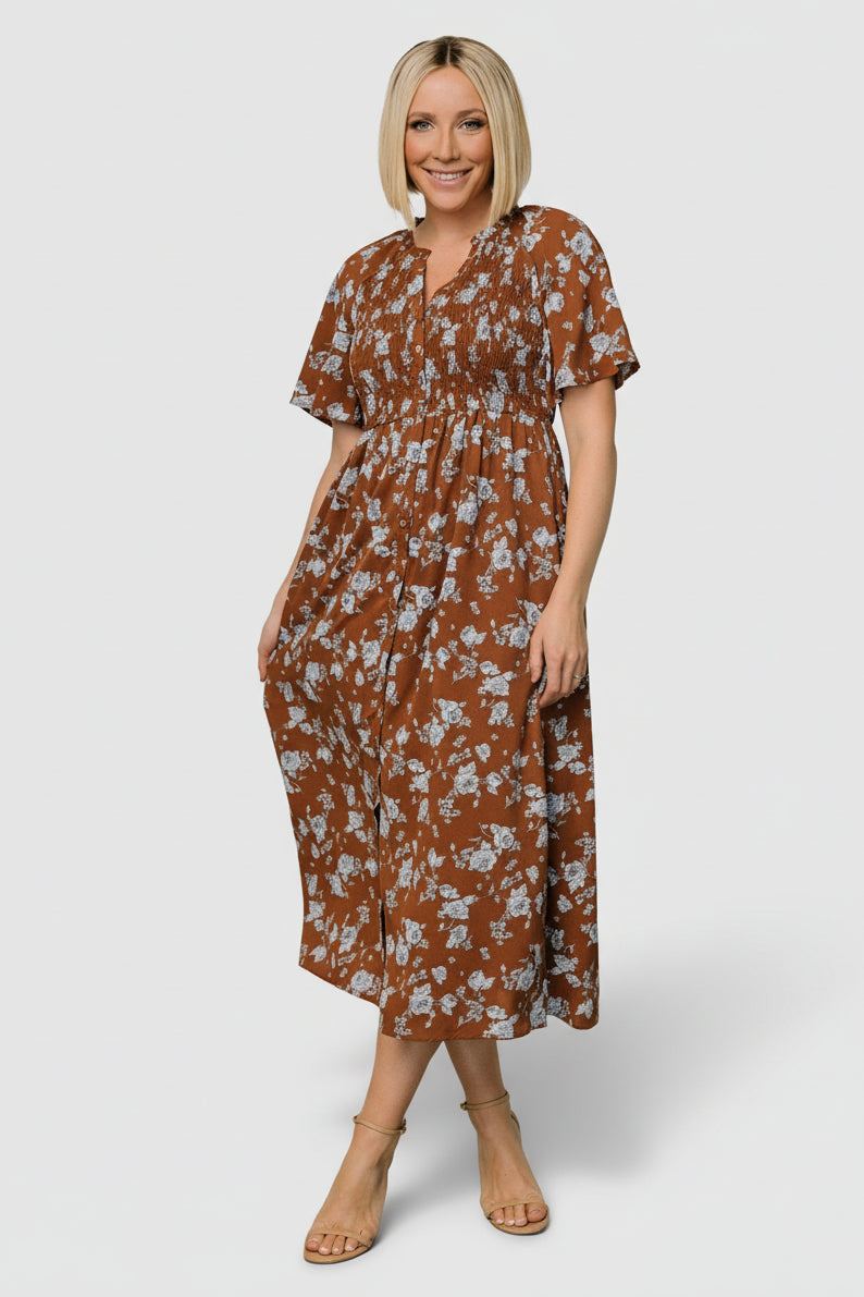 Front view of brown midi dress with blue floral print, smocked front bodice, ruffled notched neckline, button-front closure, and midi skirt with pockets.