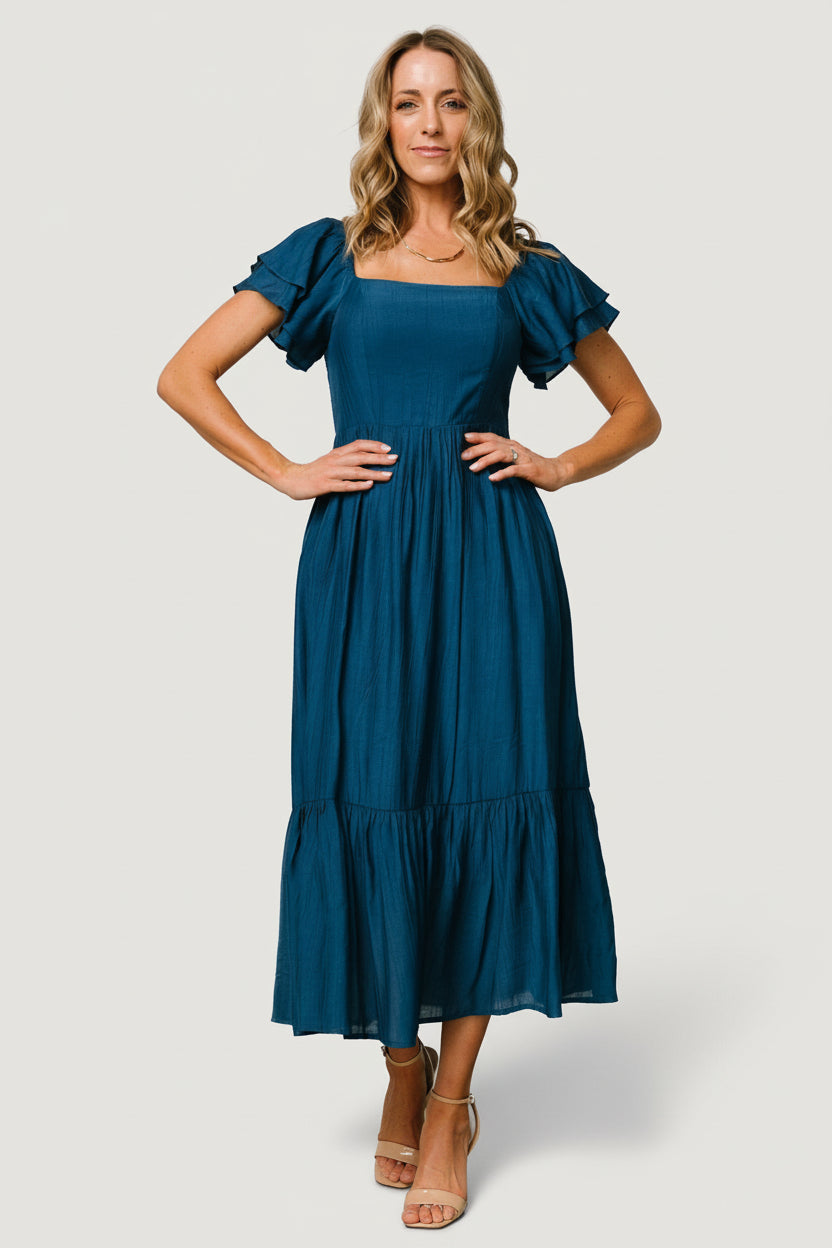 Model wearing dress styled with sandals, full-length shot showing silhouette, midi hem, and delicate ruffle details.